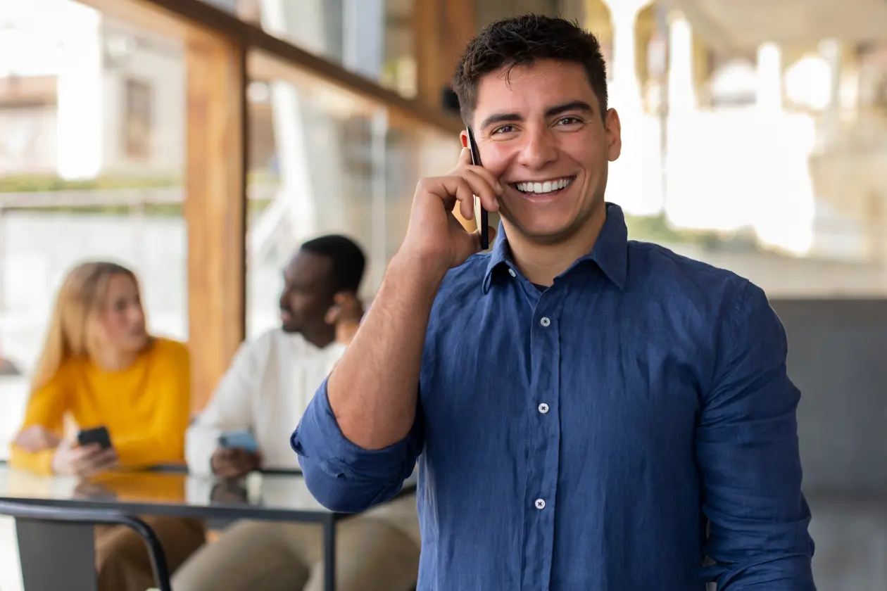 Um homem de pele clara, cabelo preto curto e usando camisa azul social sorrindo e falando ao telefone. Ao fundo, uma mulher de pele clara, com cabelo loiro solto e usando blusa amarela de manga longa, e um homem afro descendente usando camisa branca de manga longa, sentados à mesa conversando.