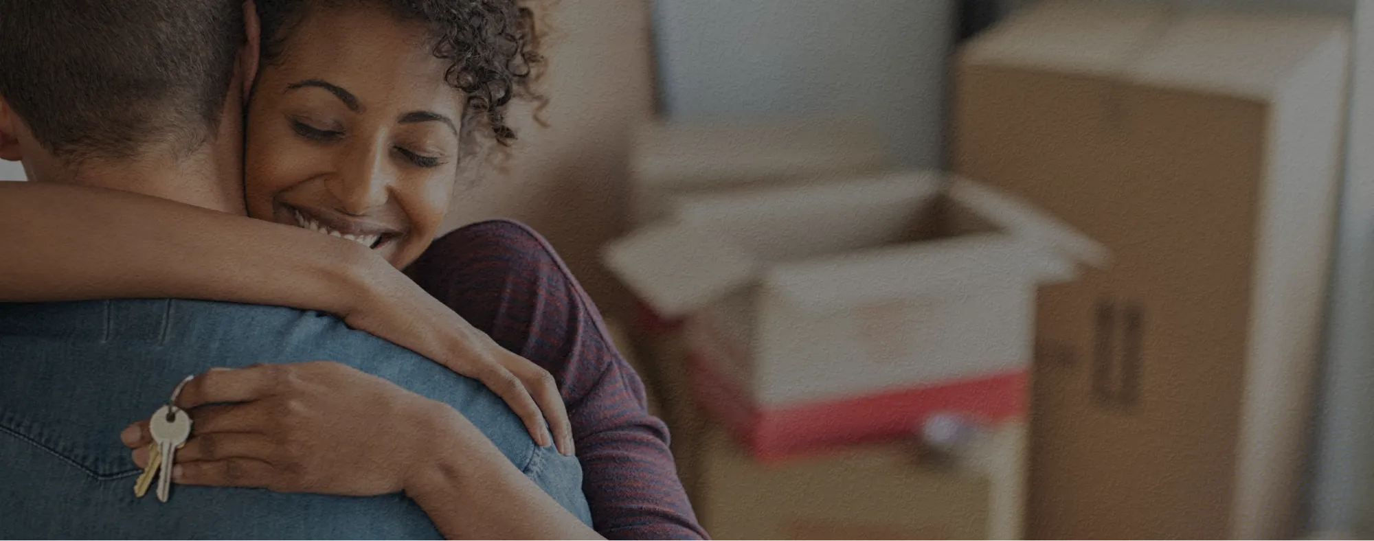 Mulher afro-descendente, de cabelo preso e usando camisa roxa com chaves na mão e abraçando um homem, que está de costas, de pele branca, cabelo curto preto, usando camisa azul. Ao fundo, caixas de papelão de mudança fechadas e abertas.