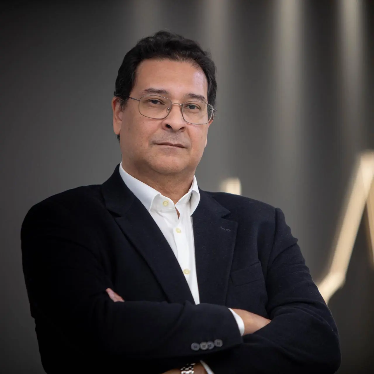 Um homem pardo, de cabelo preto, curto, usando óculos, camisa branca social e terno preto, de braços cruzados com expressão séria. Ao fundo,  uma parede cinza.
